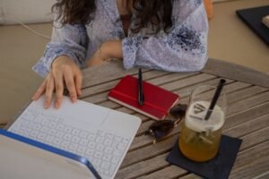 A woman works on a laptop at an outdoor café table with a drink, notebook, and sunglasses.