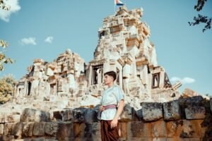 Young man in traditional clothing stands by Cambodia's ancient ruins under a clear sky.