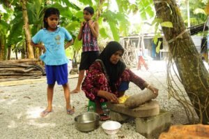 A woman preparing food outdoors in a village setting with children watching.