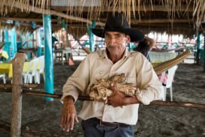 A senior man in a cowboy hat stands by a wooden fence, holding bundles in a beachside shack setting.