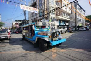 Blue jeepney driving through a vibrant city's bustling street on a sunny day.